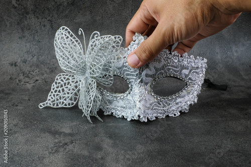 A hand holds a silver lace masquerade mask with butterfly detail on a dark textured surface. Suitable for party preparation, masquerade costume, carnival event, Halloween accessory