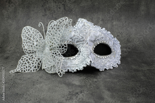 A silver lace masquerade mask with butterfly detail is placed on a dark grey textured surface. Suitable for masquerade party, mystery theme, Halloween costume, carnival event