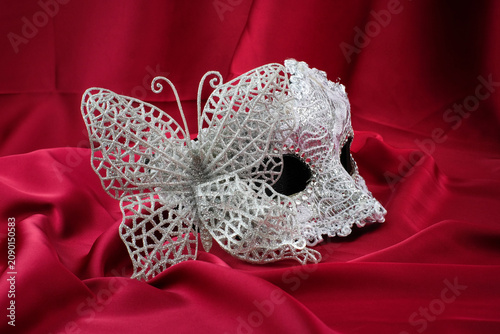 A silver decorative masquerade mask stands on red satin fabric with soft folds in the background. Suitable for masquerade party, carnival celebration, Halloween costume, theatrical event
