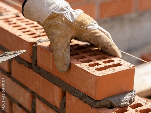 A close-up shot of a gloved hand placing a red clay brick onto a wet mortar bed while laying a wall.