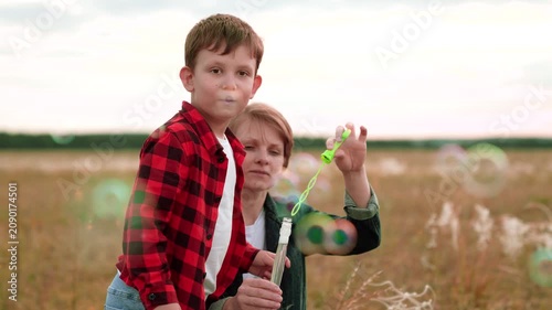 Boy with mother in grassy field blowing bubbles creating source of fun. Mom with boy enjoys activity with smiles on faces. Mum and son spend time with positive emotions making cherished memories