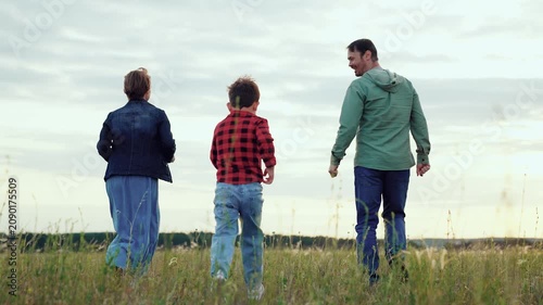 Happy family runs along grassy meadow with growing plants in agricultural area. Family of parents and little son enjoys moment of spending time together. Family showing love and care to each other