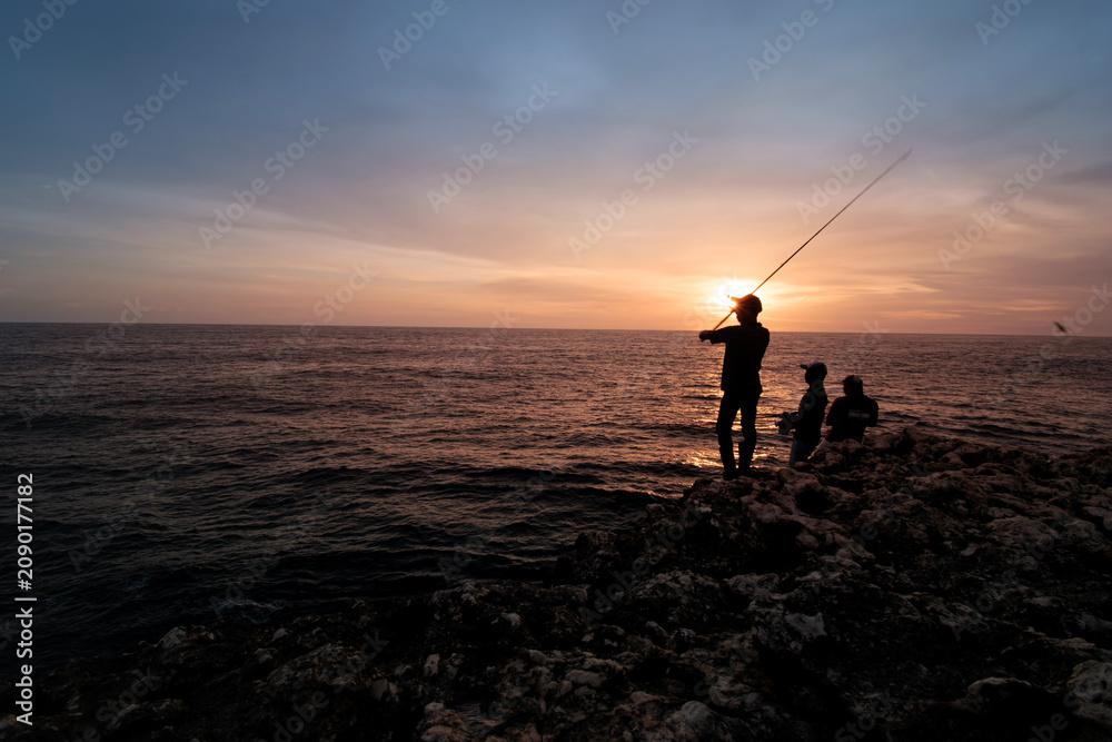 Fototapeta premium Silhouette of Fishermen Fishing on Rocky Coast at Sunset with Dramatic Ocean View, Sunset Fishing on Rocky Shore with Silhouette Anglers