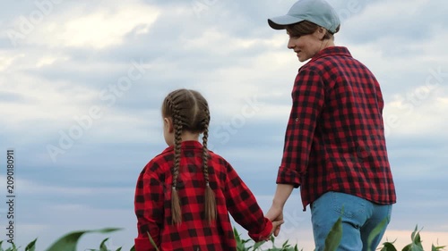 Mother agronomist talks to child holding hands on corn plantation. Mom agronomist shares moments with girl wandering on field. Daughter spends holidays with mum farmer in corn field under cloudy sky