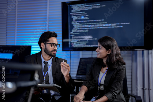 Bearded Indian engineer and South Asian businesswoman in animated discussion debating model architecture beside monitors streaming program code in a dark artificial intelligence office.