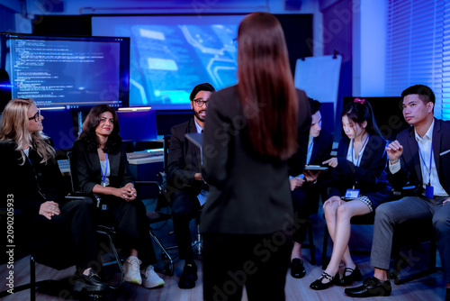 Fényképezés Businesswoman with long dark hair presenting to her team viewed from behind commanding the room's attention molecular graphics glowing on screens in the dark office on the job