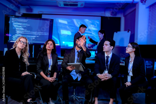 Photography Seated project team with tablets listening to a colleague at the screen murmuring agreement in a dark blue technology meeting room captured in an authentic working moment