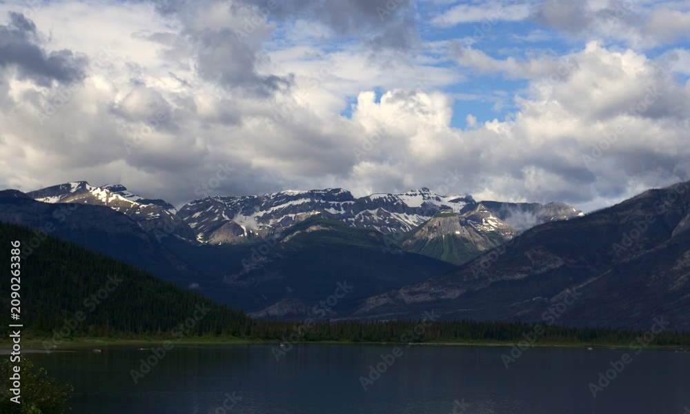 Fototapeta premium Snow-Capped Ridge Reflection: A stunning panoramic view of a massive alpine range covered in summer snow, towering over a calm lake and dense green forests under a dynamic, cloud-filled sky.