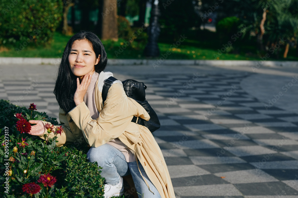Naklejka premium Woman crouching beside blooming flowers in a park, enjoying a sunny day, casual fashion, backpack ready for a stroll, candid moment of calm and beauty.