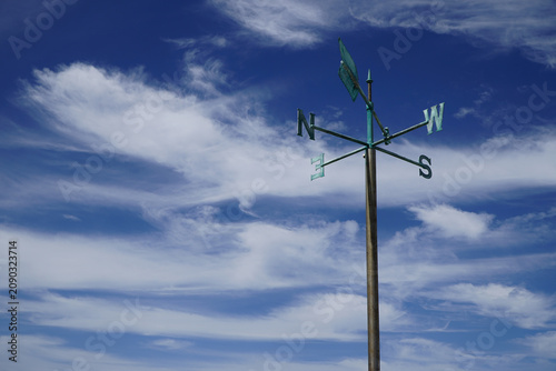 Antique copper weathervane pointing compass directions against blue sky with clouds