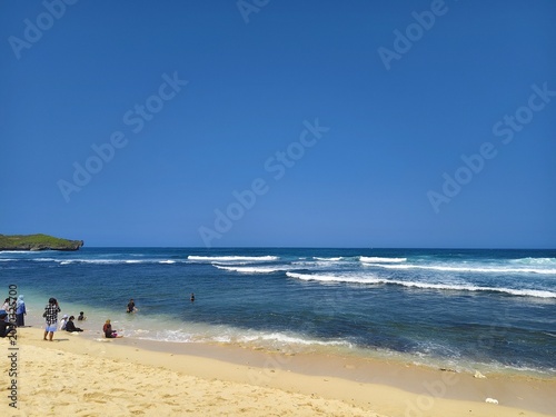 Tourists enjoying shallow ocean waters at a tropical beach with green hills under a bright blue sky