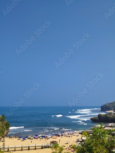 Tourists enjoying shallow ocean waters at a tropical beach with green hills under a bright blue sky
