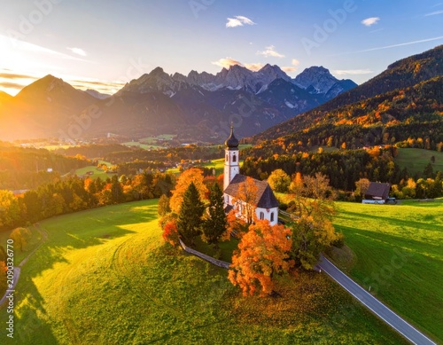 A picturesque church stands on a hill amidst autumn foliage, with majestic mountains in the background under the warm glow of sunset.