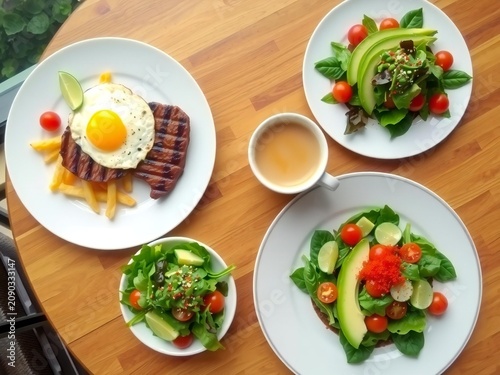 A high angle view of a wooden table set with steak, fried eggs, fresh salads and a cup of coffee
