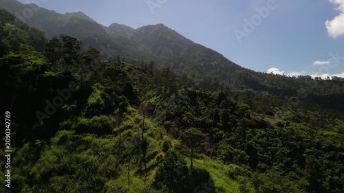 4k aerial drone video footage of Mount Merapi volcano seen from Klangon Hills emitting volcanic smoke from its summit crater, surrounded by forest mountain ridges, clear blue daytime sky.