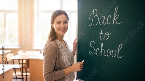 Teacher stands beside a chalkboard written with the phrase Back to School while smiling at the camera in a brightly lit classroom setting during the start of the new academic school year term.