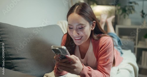 Happy Asian woman smiling while lying on sofa and using smartphone for social media application at home at night.