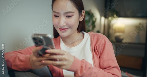 Beautiful Asian female holding smart phone, playing social media while online messaging in living room at night.