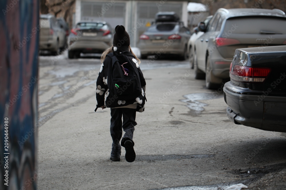 Fototapeta premium Young girl with a school backpack walking away down a city street alley between parked cars, back view.