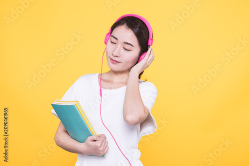 Young woman with books listening to music on yellow background
