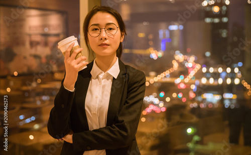 Young businesswoman with takeaway coffee in cafe at night by city lights