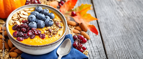 Pumpkin smoothie bowl with blueberries and granola on a rustic wooden table with autumn leaves and copy space