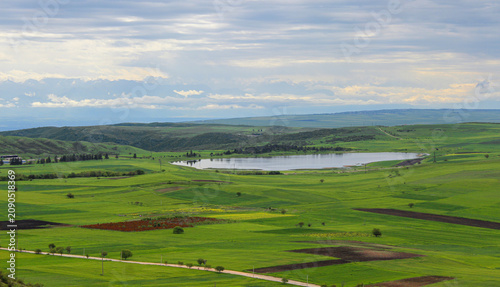 Green, plowed fields among hills covered with grass and shrubs. An artificial lake. The Caucasus Mountains. A blue sky with clouds in the background. Kakheti, Georgia