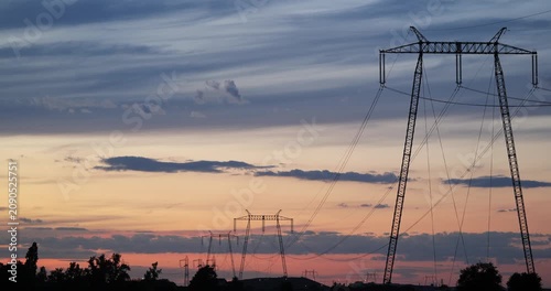 High voltage power lines and electricity pylons silhouette against a dramatic orange sunset sky in the evening