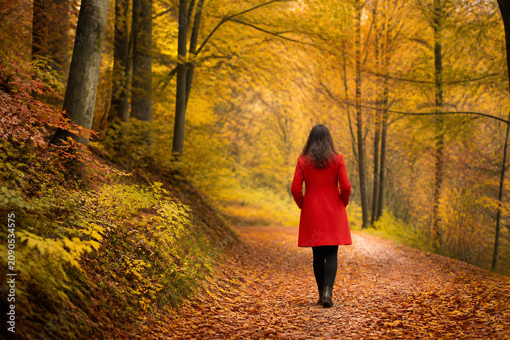 Naklejka premium A woman in a red coat walks down a path covered in autumn leaves surrounded by yellow and orange trees.