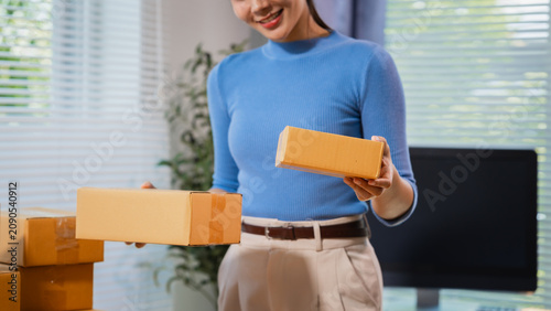 Fotografía Smiling young woman in a home office holding cardboard parcels, showing online shopping, small business shipping, packing orders, and home delivery work