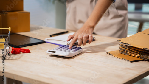 Close view of a small business worker using a calculator at a packing table with boxes, envelopes and a laptop, showing shipping costs, budgeting and online order work.