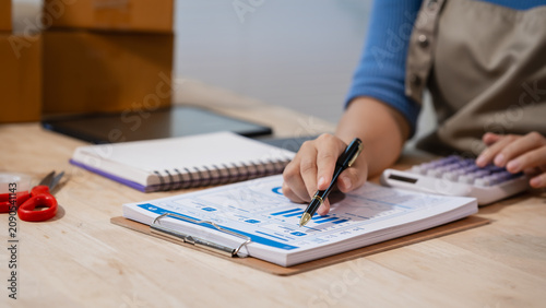 Person writing on a clipboard form while using a calculator at a desk, with notebook and boxes nearby, showing small business bookkeeping, budgeting, and home office planning.