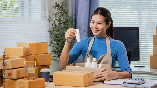 Young woman packing beauty products in cardboard boxes at a home office, checking a label for online orders. Clean workspace suggests small business, shipping, retail, and entrepreneurship.