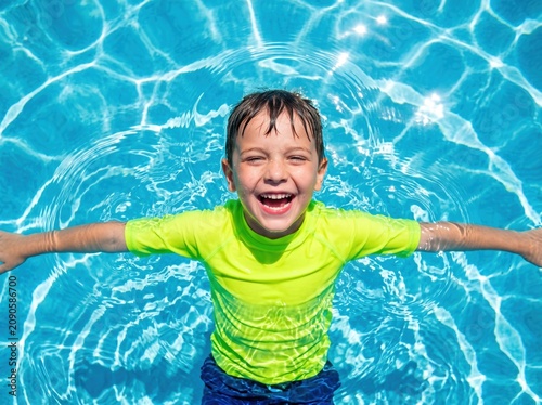 Smiling boy in bright swim shirt floating on his back in clear swimming pool water, arms outstretched, wet hair, sunlit ripples and reflections around him