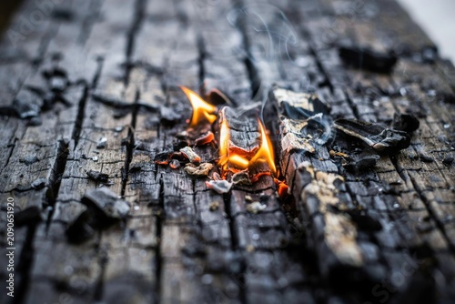 Small flames flickering on charred black wood surface with wisps of smoke rising from dark embers.