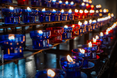 Rows of burning unlit red and blue votive candles on a stand. High angle close-up shot of white wax and wicks in glass cups, creating a repeating pattern.