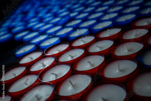 Rows of unlit red and blue votive candles on a stand. High angle close-up shot of white wax and wicks in glass cups, creating a repeating pattern.
