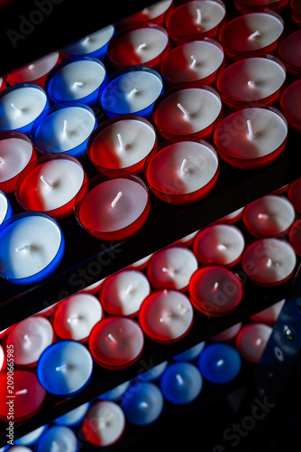 Rows of unlit red and blue votive candles on a stand. High angle close-up shot of white wax and wicks in glass cups, creating a repeating pattern.