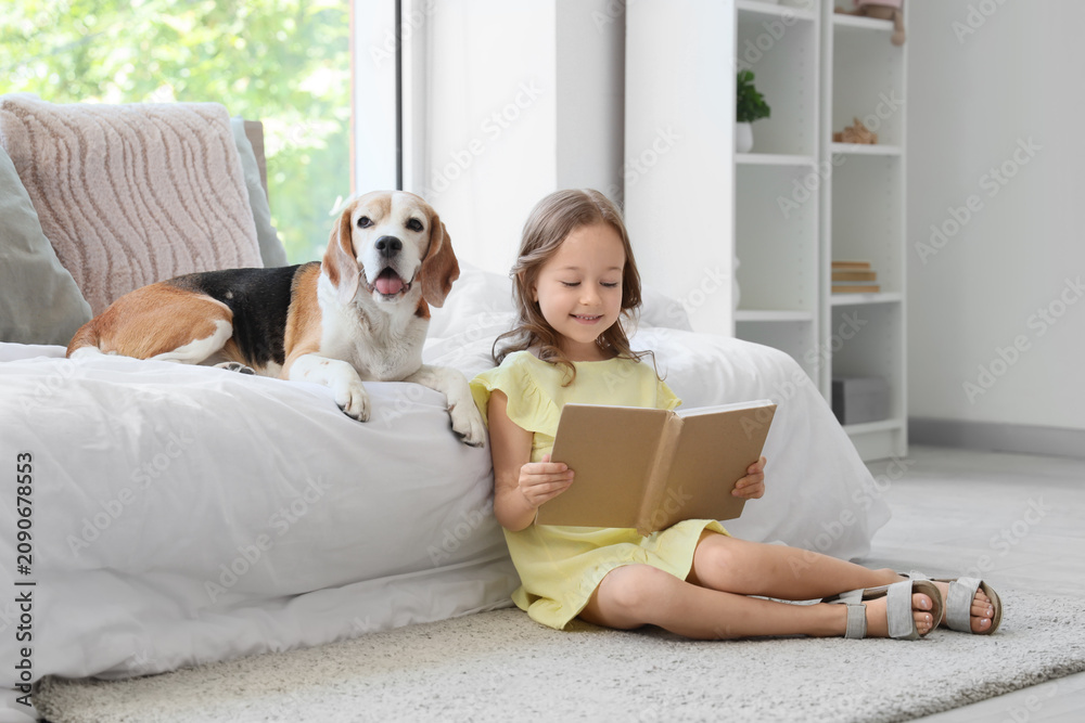 Obraz premium Cute little girl with Beagle dog reading book near bed in bedroom