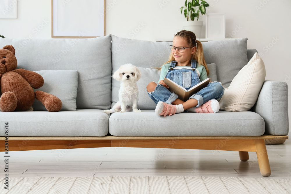 Obraz premium Cute little happy girl with Maltese dog reading book on sofa at home