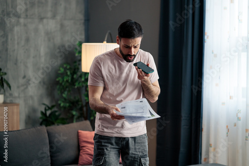 Bild auf Leinwand Young man experiencing financial stress holding bills and smartphone