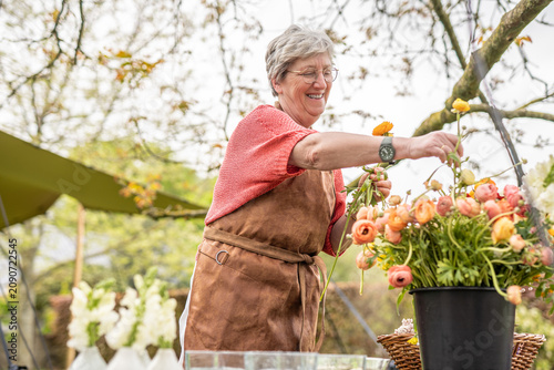 A cheerful senior woman in a brown apron smiling while arranging a colorful bouquet of ranunculus flowers at an outdoor table, celebrating creativity and joy in floristry.