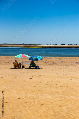 Two people relaxing in chairs under colorful umbrellas on a vast golden sand beach during a bright sunny summer day in Ile de Re, France.