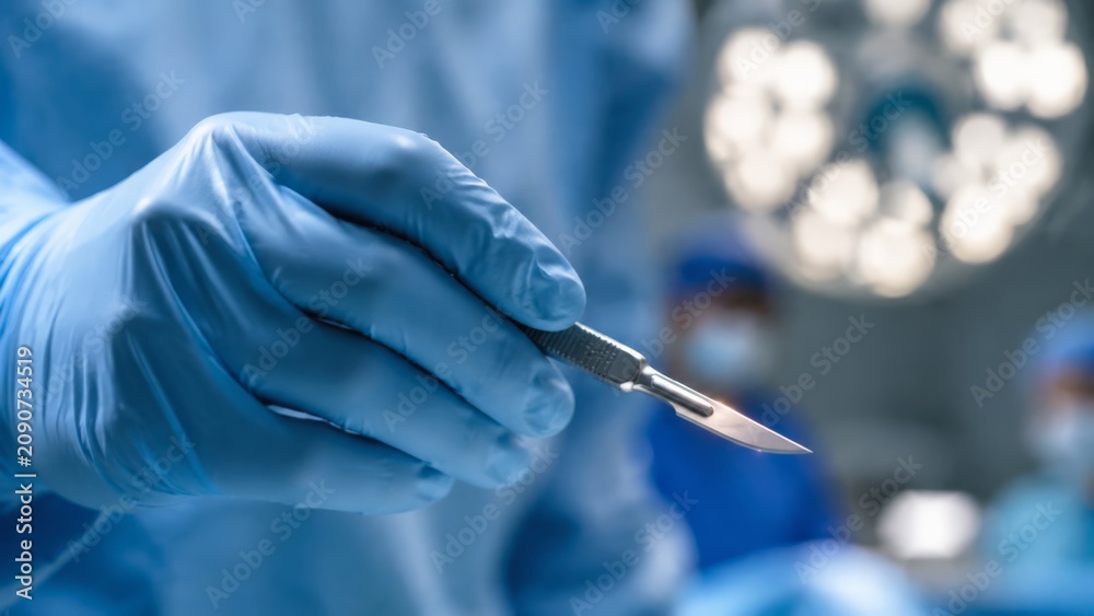 Fototapeta premium Close-up shows blue sterile glove gripping sharp scalpel beneath blurred round operating lights, with cool hospital tones, precise medical focus, and clinical atmosphere for surgery concepts