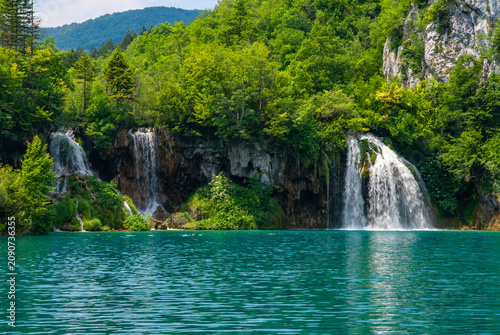 Vibrant turquoise lake mirrors lush green forest, with multiple cascading waterfalls flowing down rugged cliffs, creating breathtaking natural landscape. Plitvice Lakes is National Park in Croatia.