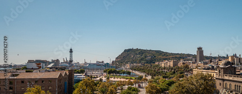 A panoramic view of Barcelona featuring the coastline, hills, and urban architecture under a clear blue sky. The scene captures the vibrant city life and natural beauty.