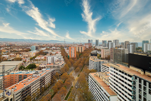 Aerial view of Diagonal Mar district in Barcelona showcasing modern buildings, tree-lined streets, and a clear blue sky with wispy clouds. The cityscape reflects urban life and architecture.