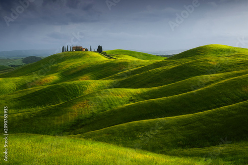The Podere Staffolino sits on a rounded hilltop surrounded by deeply saturated green rolling wheat fields, a distant rain shower visible under a stormy sky. Crete Senesi, Tuscany, Italy.