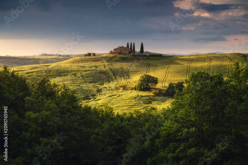 A stone farmhouse with three flanking cypresses sits spotlit on a bright green ridge, dense dark woodland framing the glowing hillside below. Val d'Orcia, Tuscany, Italy.