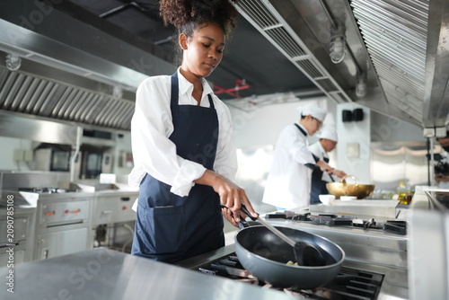 Quadro su tela Young female culinary student cooking holds the pan handle while sauteing in the